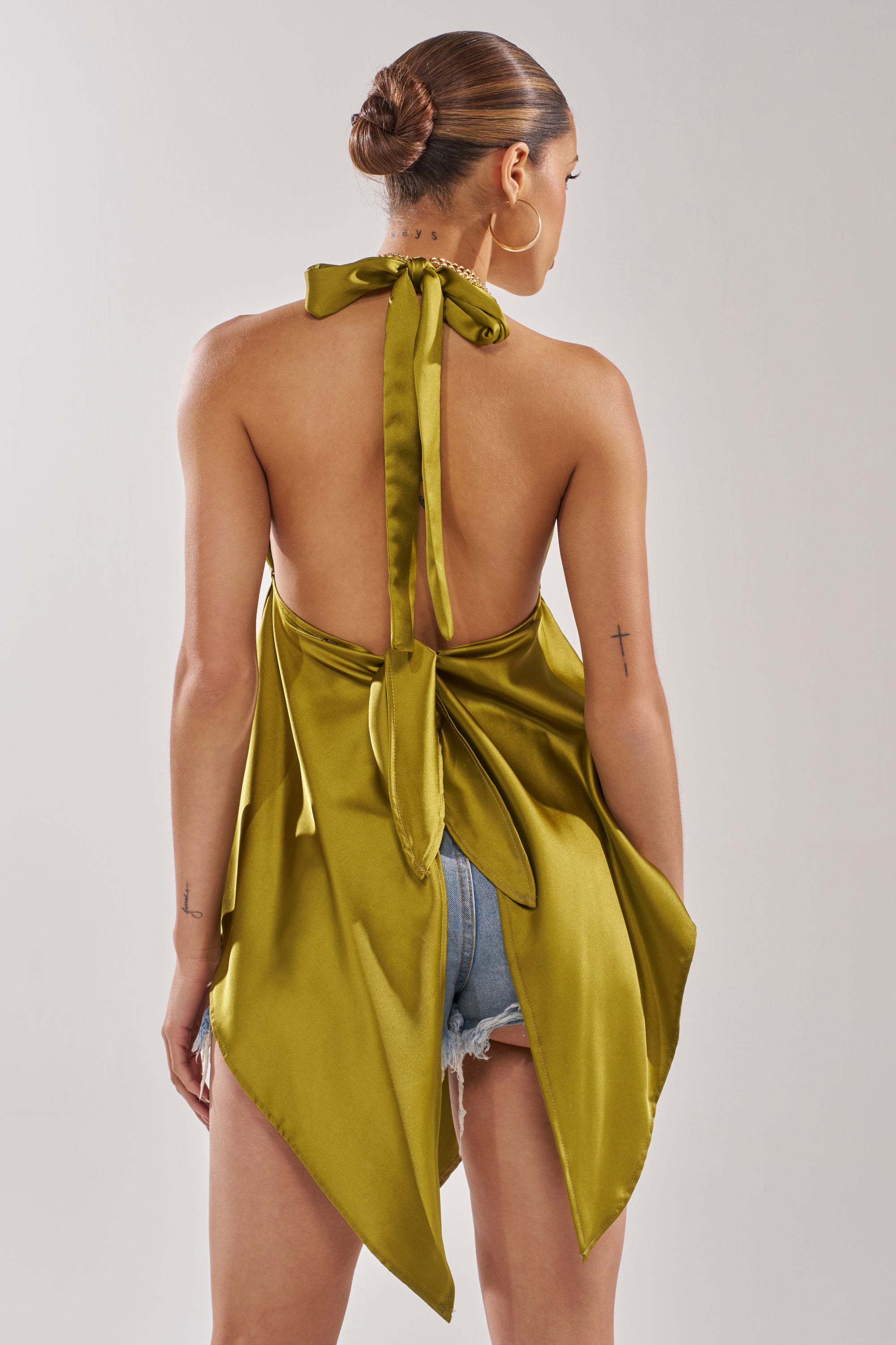 A woman with a sleek bun wears the LOTS OF LOVE TO GIVE HALTER NECK BLOUSE IN CHARTREUSE— a backless satin halter top with a draped handkerchief hem, tied in a bow and styled with light denim shorts to highlight its open back and flowing silhouette. 5