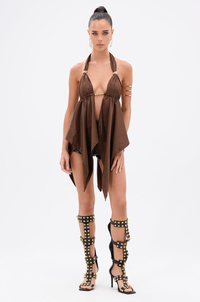 A woman stands against a white background wearing the LOTS OF LOVE TO GIVE HALTER NECK BLOUSE IN BROWN, paired with black shorts, gold arm cuffs, and tall black gladiator sandals featuring gold-tone ring accents.