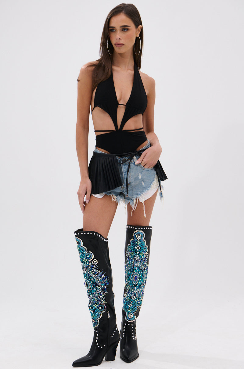 A woman poses in the SHOW IT OFF CUT-OUT BODYSUIT IN BLACK, paired with denim shorts and tall black boots featuring turquoise and silver embroidery, set against a plain white background. 4