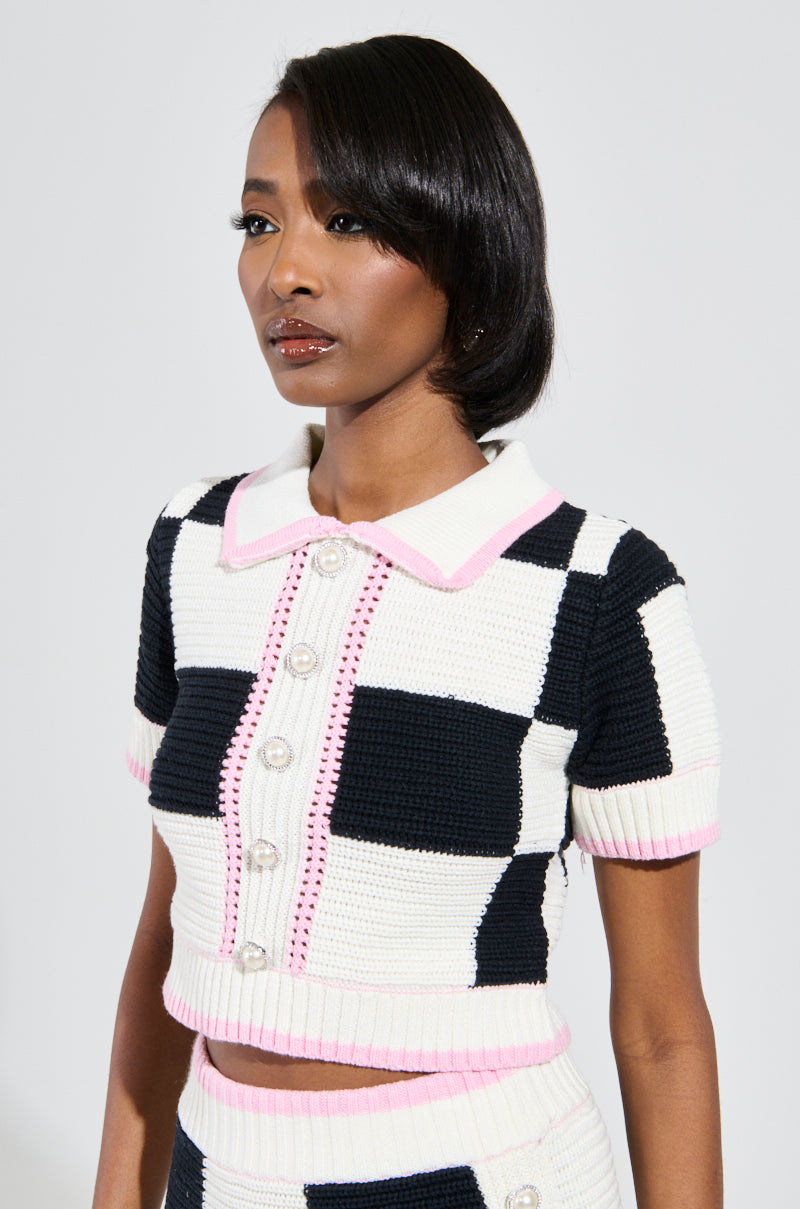 A woman wears the HIGH SPIRITS SHORT SLEEVE BUTTON DOWN SWEATER, showcasing a stretchy knit design with a black and white color block checkered print accentuated by pink details. The sweater features short sleeves, a collar, and front buttons. Her straight hair cascades to one side as she maintains a neutral pose against a plain background.