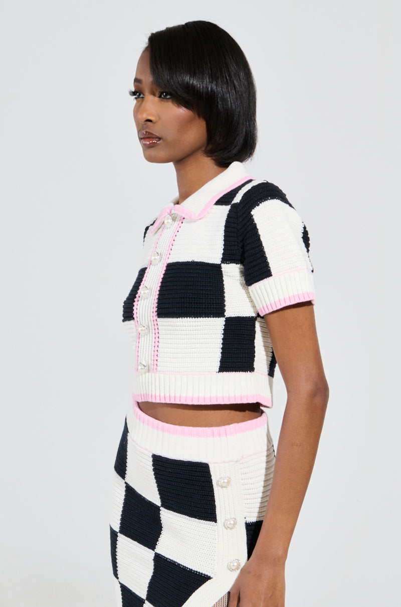 Dressed in the HIGH SPIRITS SHORT SLEEVE BUTTON DOWN SWEATER, featuring a black and white color block checkered print with pink trim, the ensemble includes a cropped top paired with a matching skirt. Set against a plain white background, the person gazes thoughtfully to the side.