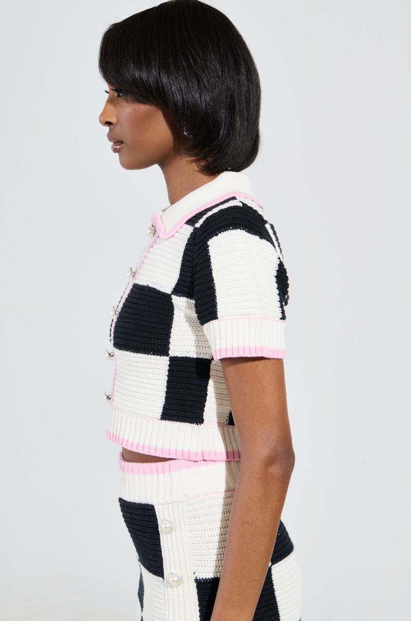 A person with short hair is wearing the High Spirits Short Sleeve Button Down Sweater, a black and white color block checkered print knit top and skirt ensemble adorned with pink trim. The outfit boasts a chic cropped silhouette and stretchy knit fabric, as they stand in profile against a plain background.