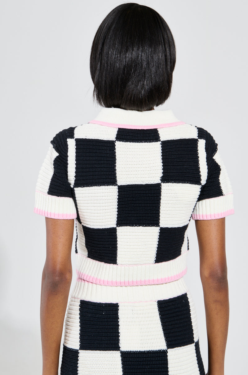 A person facing away from the camera, flaunting the HIGH SPIRITS SHORT SLEEVE BUTTON DOWN SWEATER, a stretchy knit ensemble with a cropped silhouette. The black and white color block checkered print is accented by pink trim on both the top and skirt, all set against a plain white background.