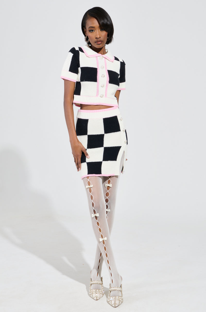 A woman poses against a plain background, wearing the HIGH SPIRITS SHORT SLEEVE BUTTON DOWN SWEATER in a chic black and white color block checkered print. Her ensemble features a stretchy knit cropped silhouette top paired with a matching skirt. She completes the look with white patterned tights, strappy high heels, and sleek short hair.