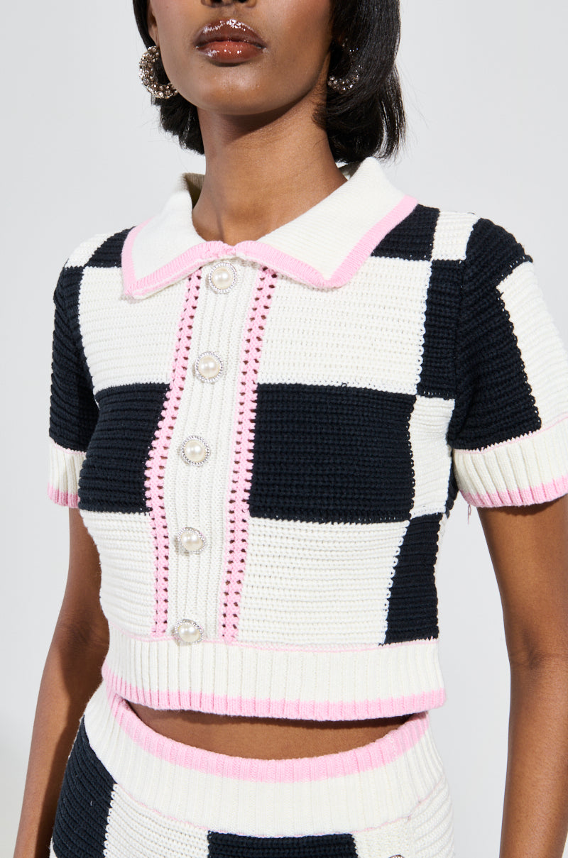 A person wears the High Spirits Short Sleeve Button Down Sweater, characterized by its knitted texture, color block checkered pattern, and chic pink trim. The top showcases a collar and pearl-like buttons down the front, creating a trendy cropped silhouette. A glimpse of the coordinating skirt is also visible.