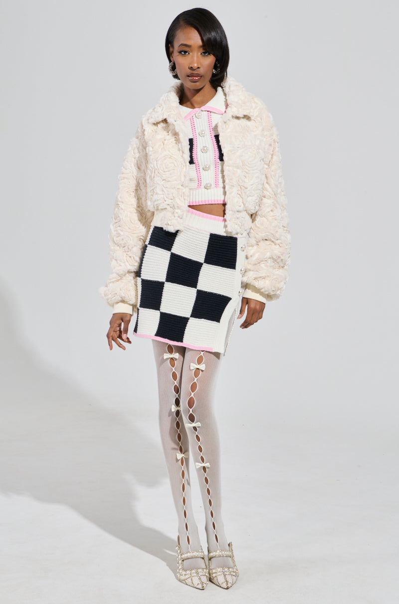 A person stands in a stylish outfit featuring a textured white jacket, pink and white top, a HIGH SPIRITS SWEATER KNIT MINI SKIRT with a checkered pattern, patterned stockings, and white embellished heels on a white background.