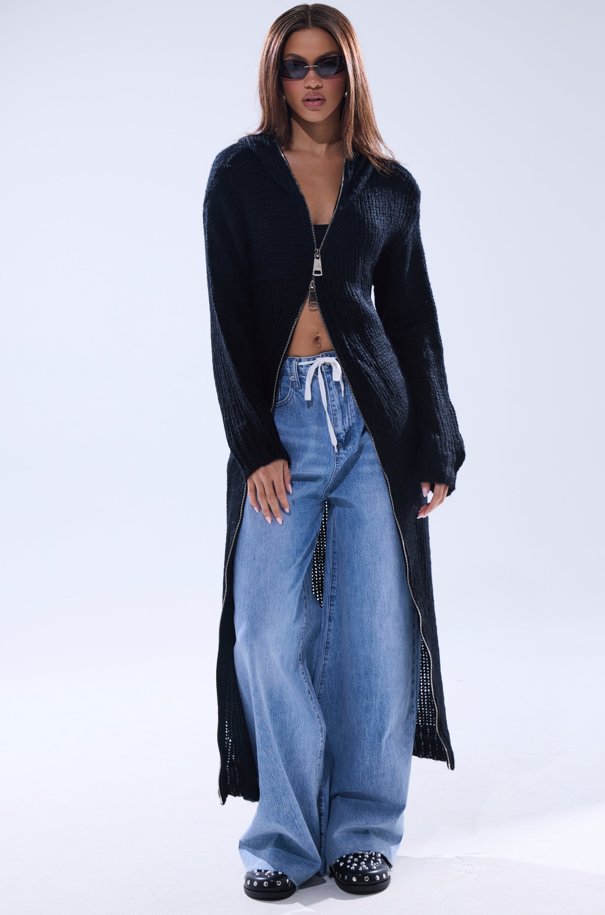 A person stands against a plain light background wearing sunglasses, the COZIEST GIRLY ZIP UP SWEATER as a black knit cardigan, a black crop top, light blue drawstring wide-leg jeans, and black studded shoes.