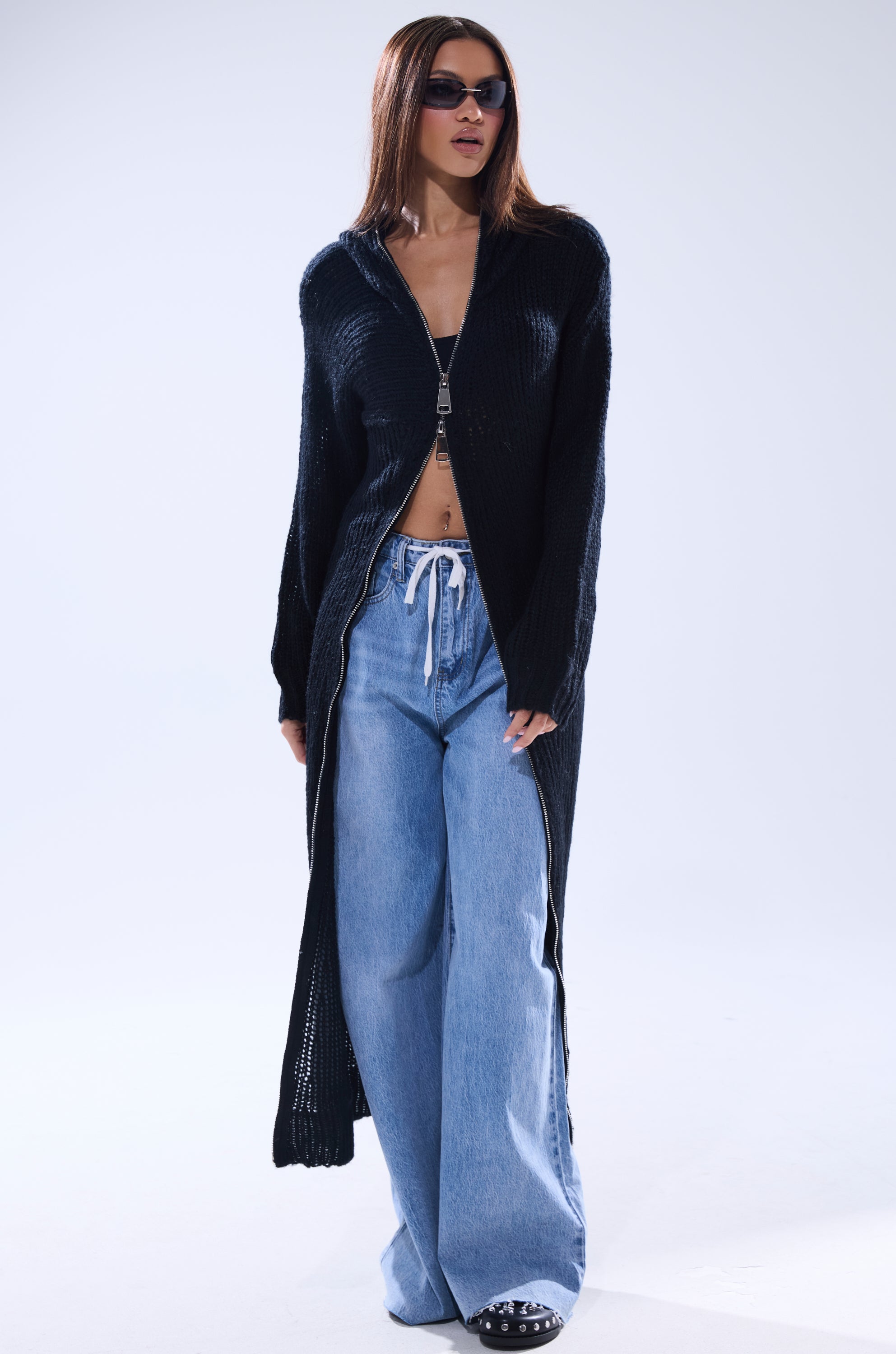 A woman stands against a plain light background wearing the COZIEST GIRLY ZIP UP SWEATER over a crop top, paired with wide-leg light blue drawstring jeans, sunglasses, and spiked black shoes.