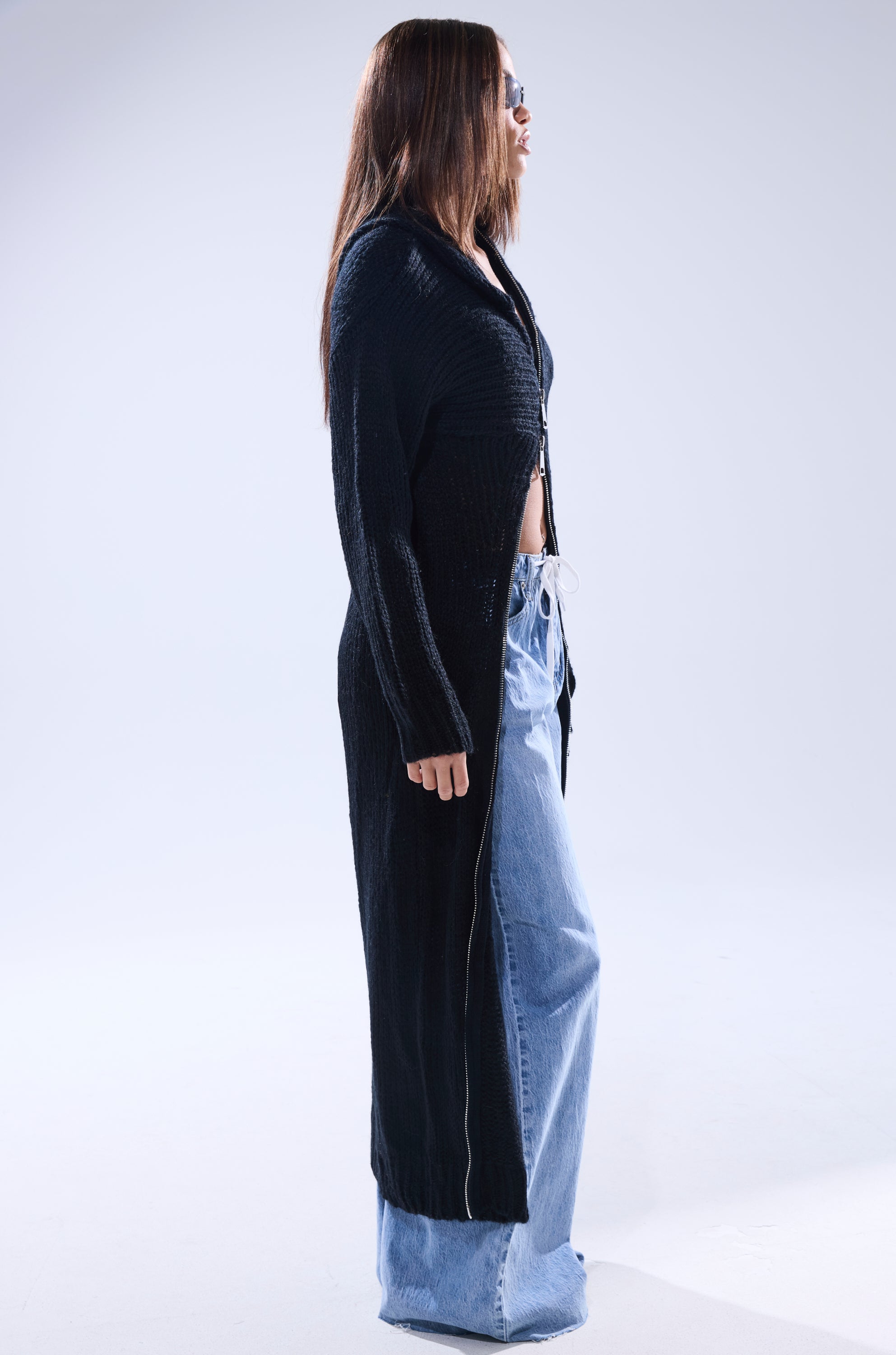 A woman stands in profile against a plain light background, wearing sunglasses, a crop top, loose light blue jeans, and the COZIEST GIRLY ZIP UP SWEATER.