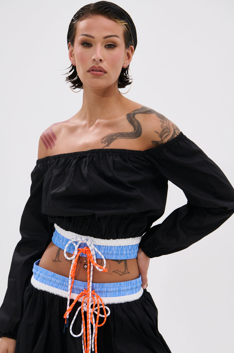 A person with short dark hair and visible tattoos confidently poses in the LET IT GO OFF THE SHOULDER BLOUSE, paired with a blue, white, and orange lace-up corset belt, against a plain light background.
