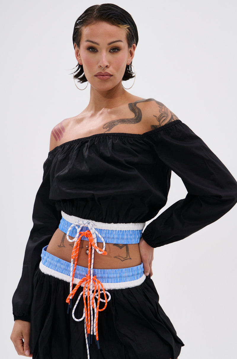 A woman with short dark hair and visible tattoos poses confidently in the LET IT GO OFF THE SHOULDER BLOUSE styled as a cropped, sporty-chic top, paired with a blue-and-white corset featuring orange laces against a plain background.