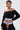 A woman with short dark hair and visible tattoos poses confidently in the LET IT GO OFF THE SHOULDER BLOUSE styled as a cropped, sporty-chic top, paired with a blue-and-white corset featuring orange laces against a plain background.