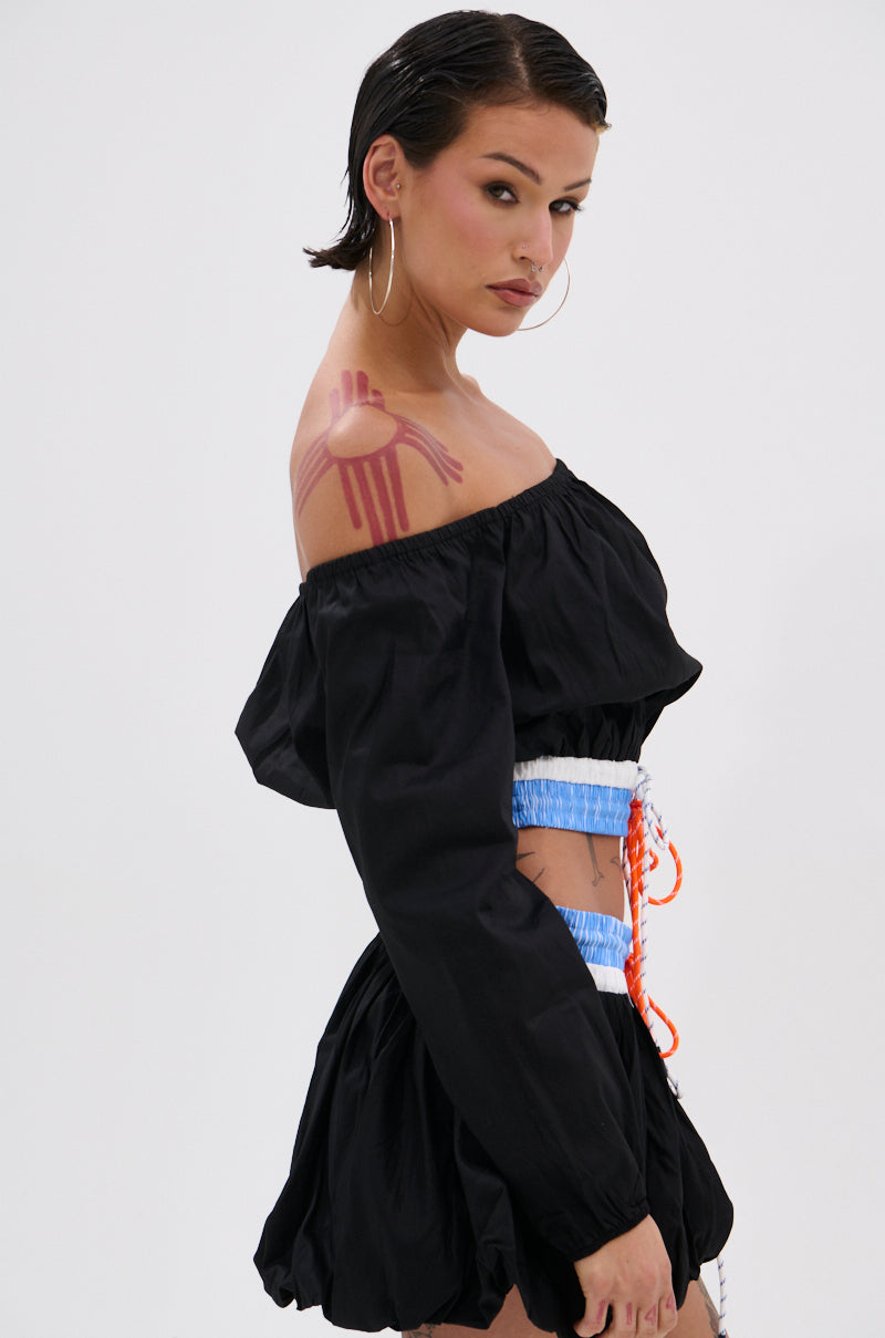 A woman with short dark hair and hoop earrings wears the LET IT GO OFF THE SHOULDER BLOUSE with blue accents, showcasing a pink and red abstract painted design on her shoulder as she gazes confidently at the camera.