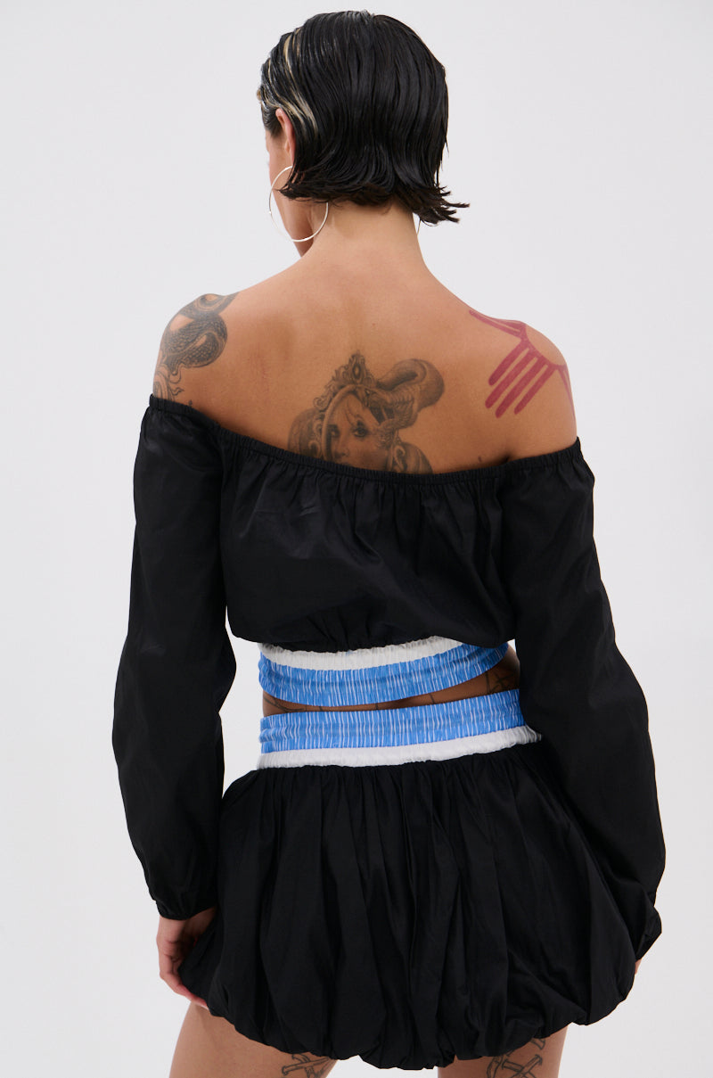 A person with short, wet-look hair faces away, wearing the LET IT GO OFF THE SHOULDER BLOUSE featuring blue and white waistband details. Tattoos—a face and pink handprint—adorn their upper back and arms.