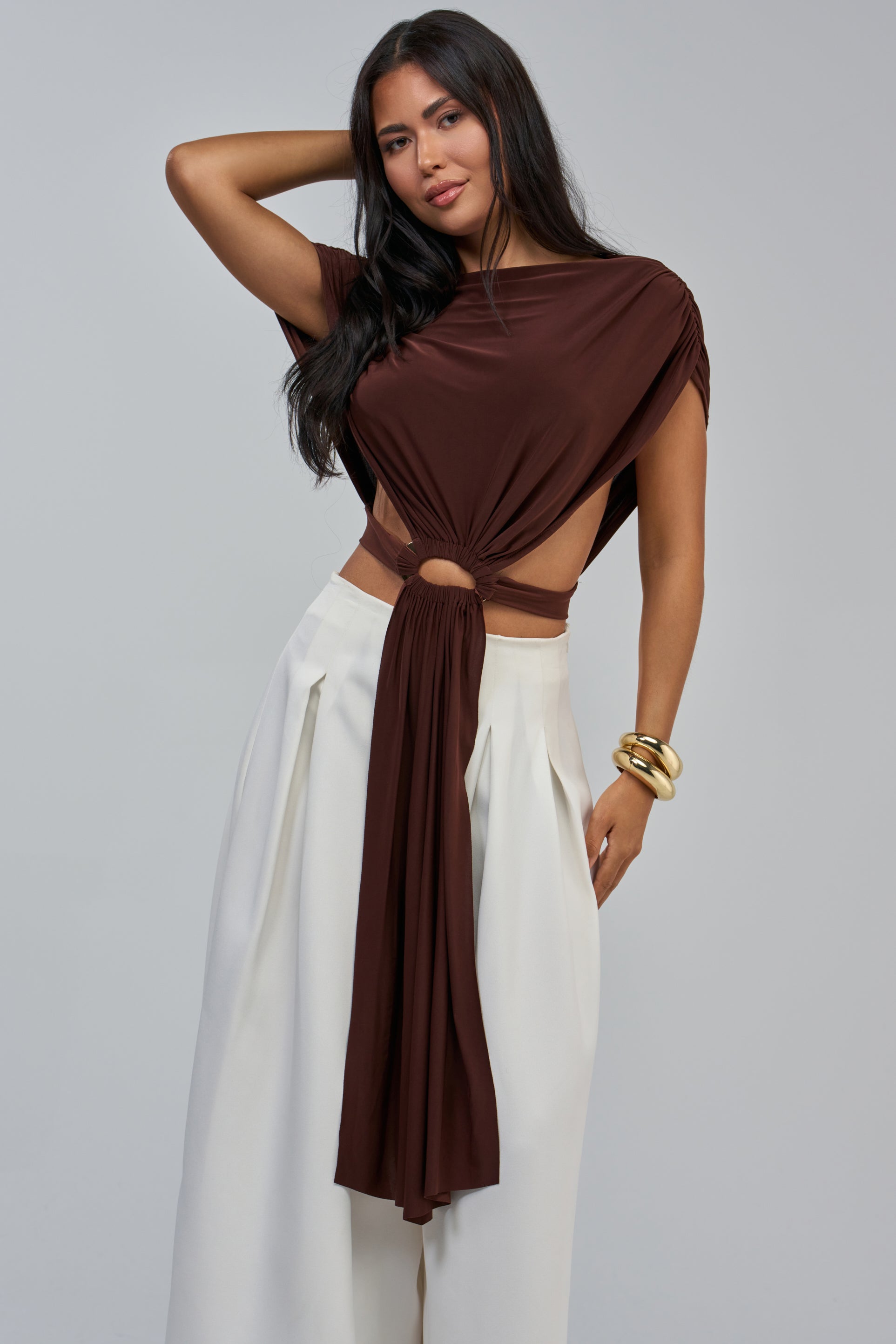 Wearing the COCOA SHIRT—a brown cropped blouse featuring a central ring accent—paired with high-waisted, wide-leg white pants, she poses with one arm raised and gold bangles on her wrist against a light gray background for a chic summer date night look.