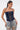A woman with dark hair in a ponytail wears the KIMMY LACE UP DENIM CORSET and light blue, distressed jeans, standing confidently against a plain white background.