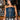 A woman with long dark hair wears the KIMMY LACE UP DENIM CORSET, styled with a concho belt, and stands before a bar lined with glassware and beer taps, gazing to the side.