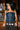 A woman with long dark hair wears the KIMMY LACE UP DENIM CORSET, styled with a concho belt, and stands before a bar lined with glassware and beer taps, gazing to the side.