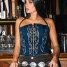 A woman with long dark hair wears the KIMMY LACE UP DENIM CORSET, styled with a concho belt, and stands before a bar lined with glassware and beer taps, gazing to the side.