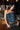 A woman with long dark hair leans against a wooden bar, wearing the KIMMY LACE UP DENIM CORSET styled cowgirl-chic with brown pants and a turquoise belt. Beer taps and glasses can be seen in the background.