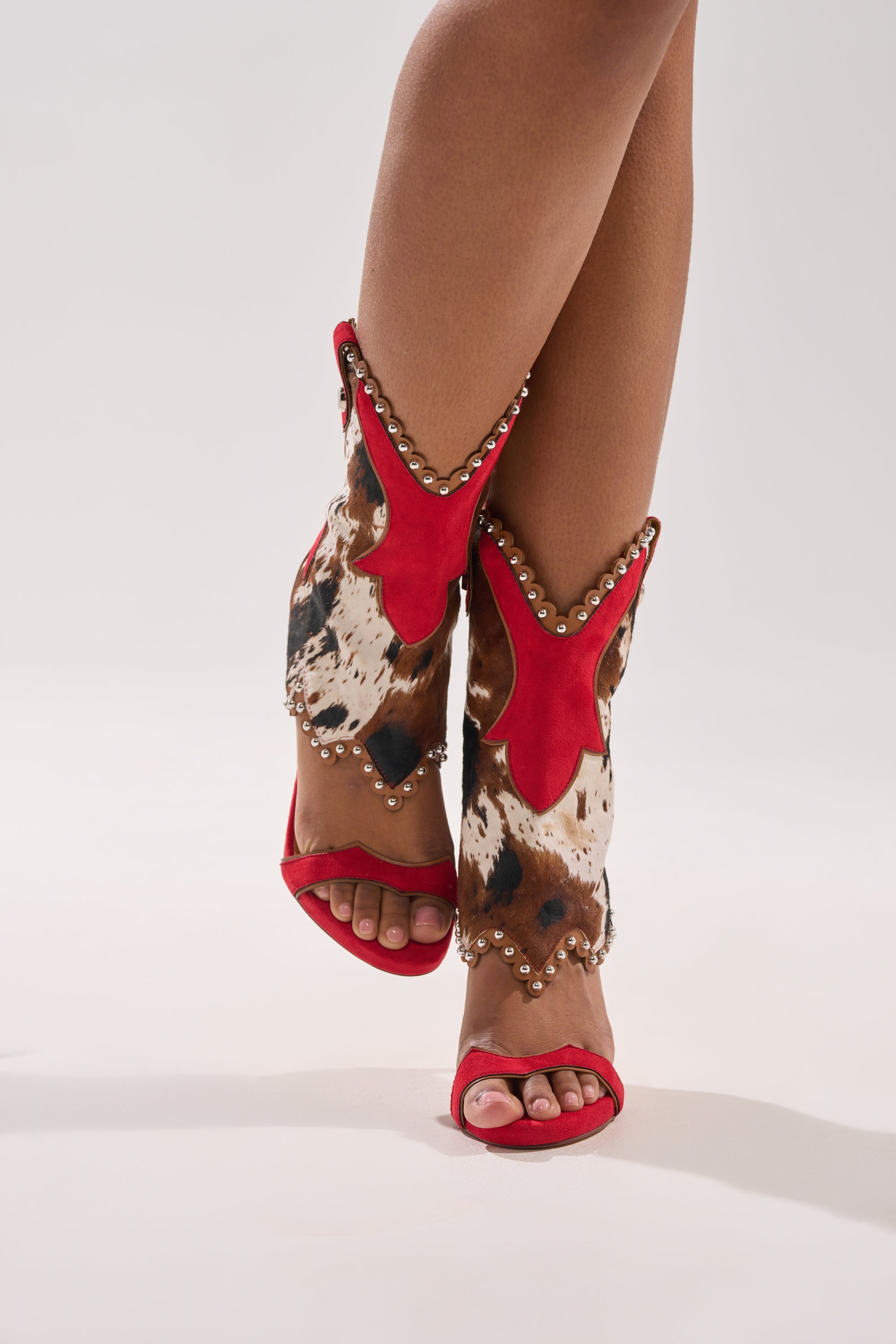 A person models the AZALEA WANG TAHLIE COW MULTI WESTERN GLADIATOR SANDAL—cow-print open-toe stiletto boots with red trim and silver studs—standing with legs crossed against a plain background. 4