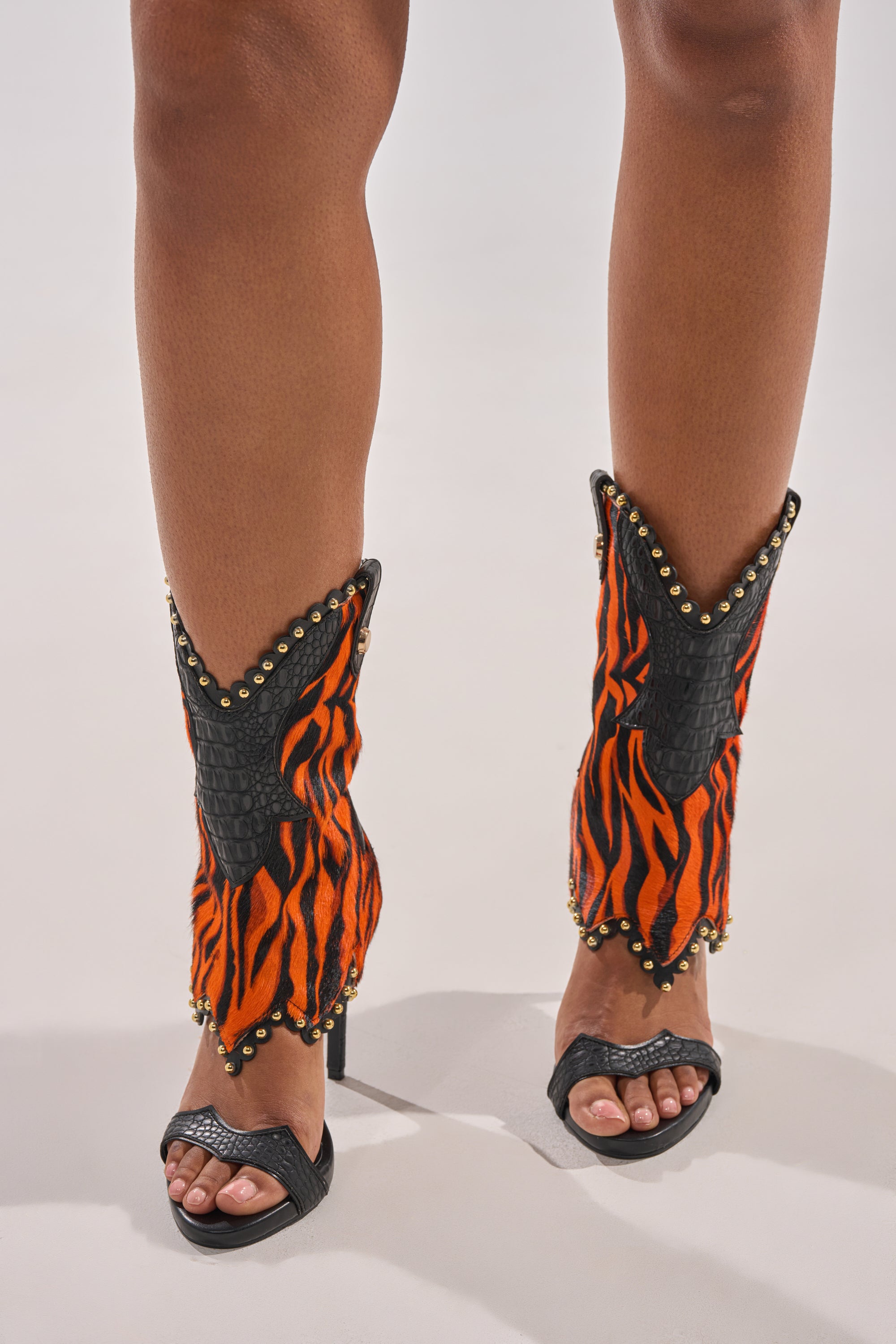 A person wears the AZALEA WANG TAHLIE ORANGE MULTI WESTERN GLADIATOR SANDAL with bold orange and black tiger-striped, gold-studded leg accessories, standing on a white background.