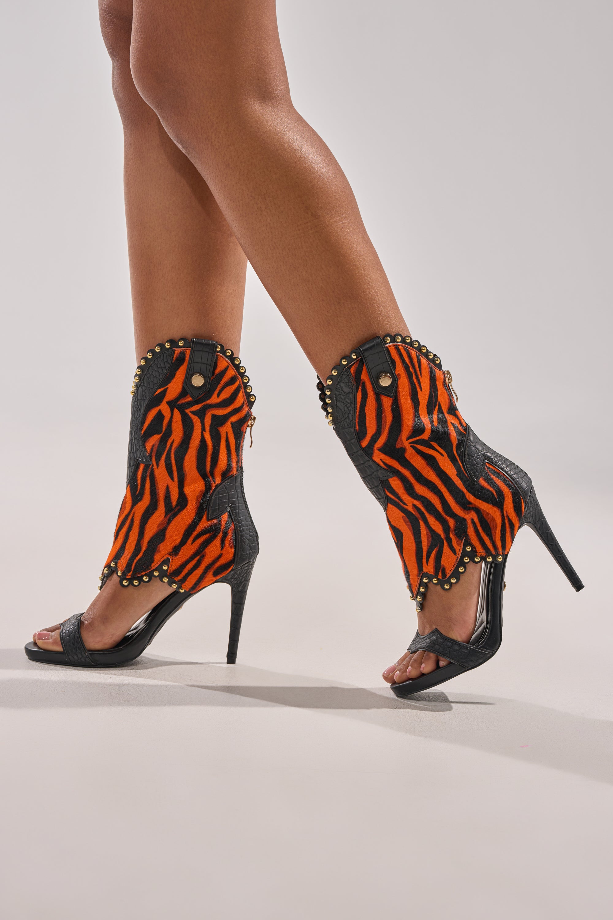 A person models the AZALEA WANG TAHLIE ORANGE MULTI WESTERN GLADIATOR SANDAL, featuring bold orange and black tiger stripes with gold studs, against a plain light background.