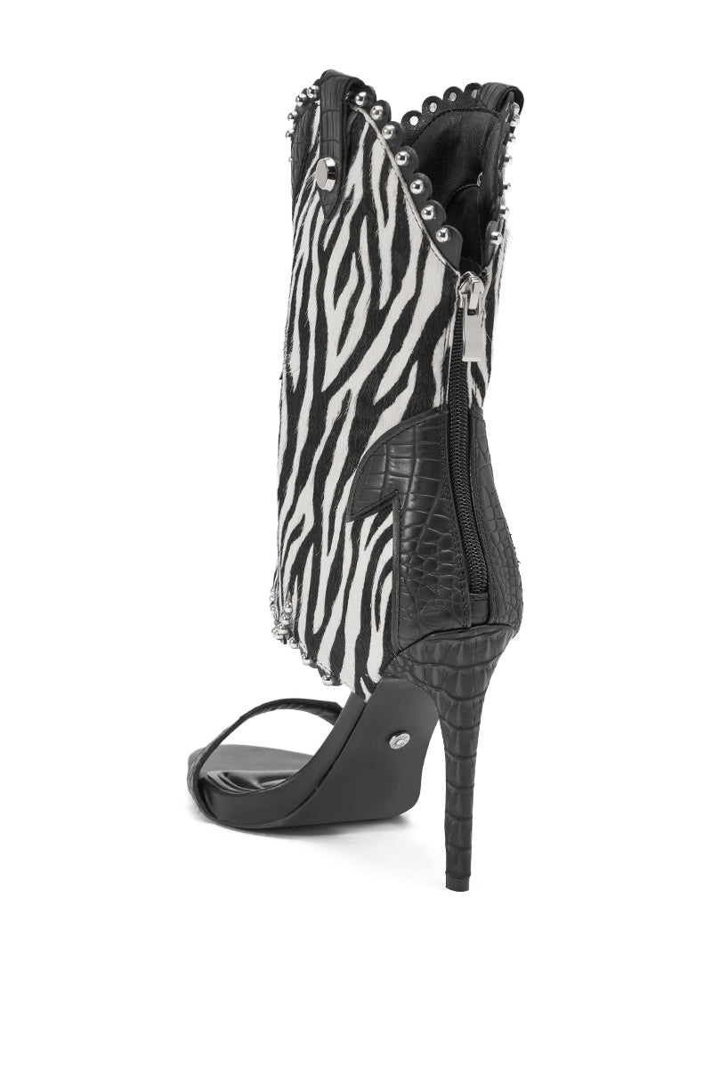 The AZALEA WANG TAHLIE ZEBRA MULTI WESTERN GLADIATOR SANDAL features a zebra-print upper, black crocodile-textured heel, edge studs, side zipper, and open toe for a bold statement look.