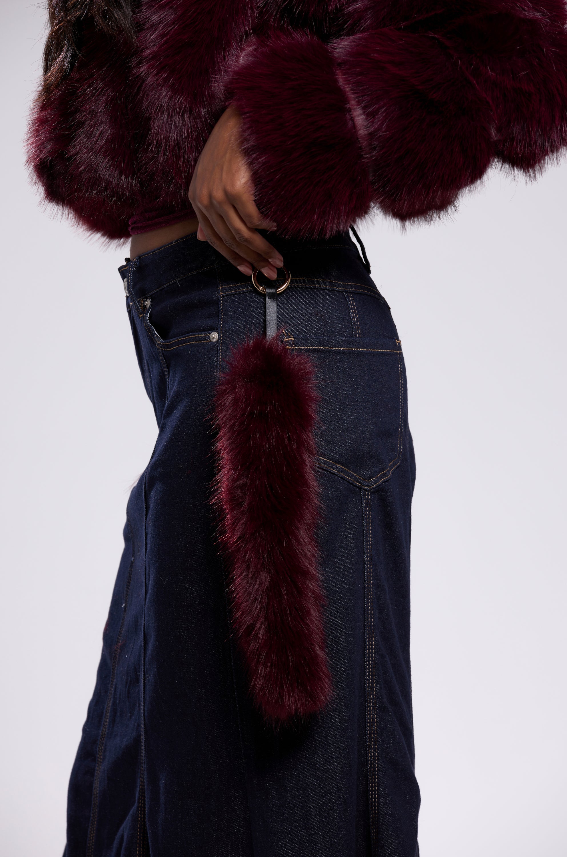 A person in a dark faux fur jacket and denim skirt holds a bag featuring the TOO GOOD FAUX FUR KEYCHAIN, a fluffy plum accessory that adds a unique touch. 0