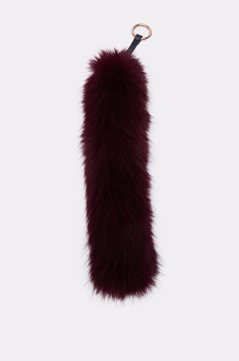 The TOO GOOD FAUX FUR KEYCHAIN features long, dark plum faux fur with a gold metal keyring atop, making it a chic bag accessory, shown on a plain light background. 1