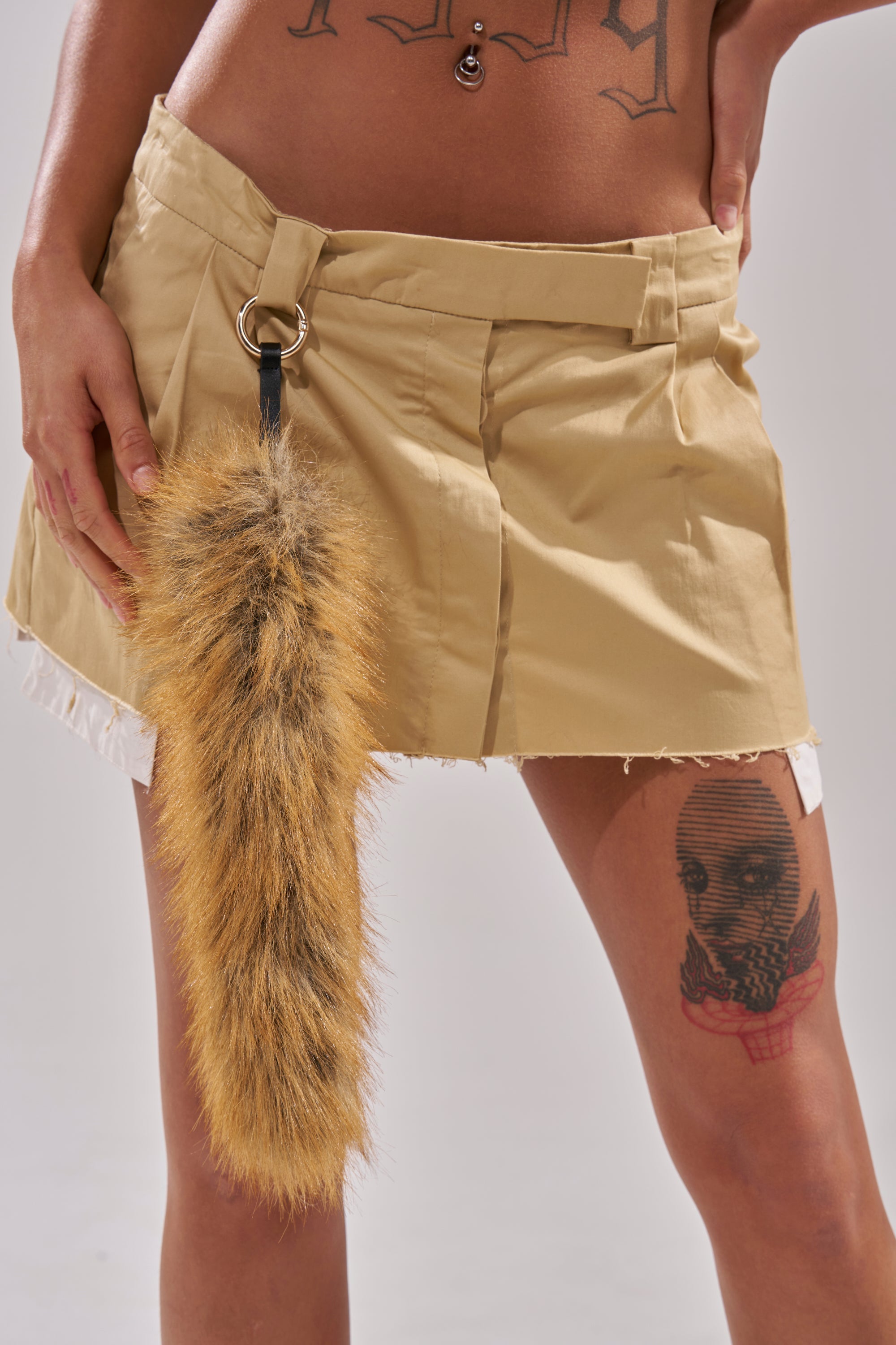 Wearing a beige mini skirt, the person sports the THAT GURL FAUX FUR KEYCHAIN—a faux animal tail with gold-tone clasp—as an accessory. Tattoos of a masked face and script adorn their torso and right thigh, with midriff exposed. 0