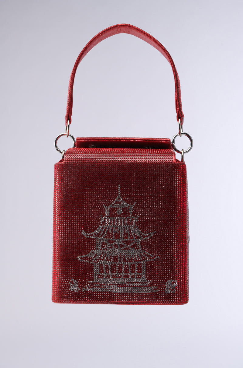 The TAKE ME AWAY BLING BAG is a red, rectangular handbag adorned with a silver embroidered pagoda and rhinestone embellishments. It has a textured look, a single handle attached to silver rings, and sits against a plain gray backdrop.