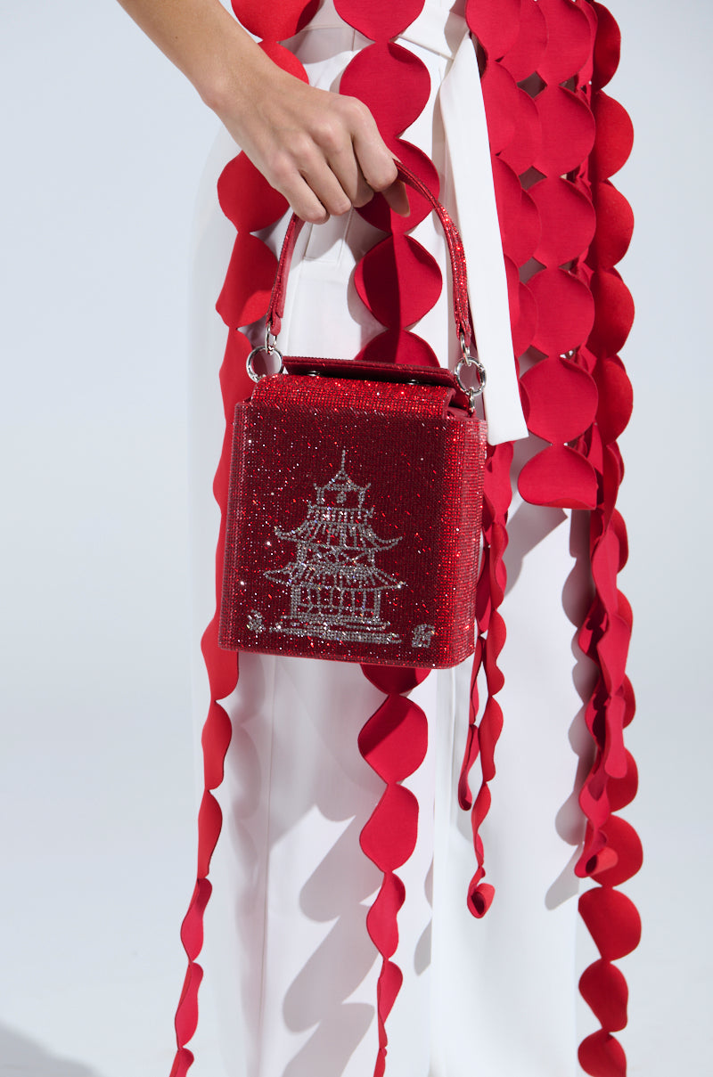 A person holds the TAKE ME AWAY BLING BAG, a red, sparkly handbag with rhinestone embellishments featuring a pagoda design. They wear white pants and a flowy red garment with circular petals in front of a softly neutral background. 1