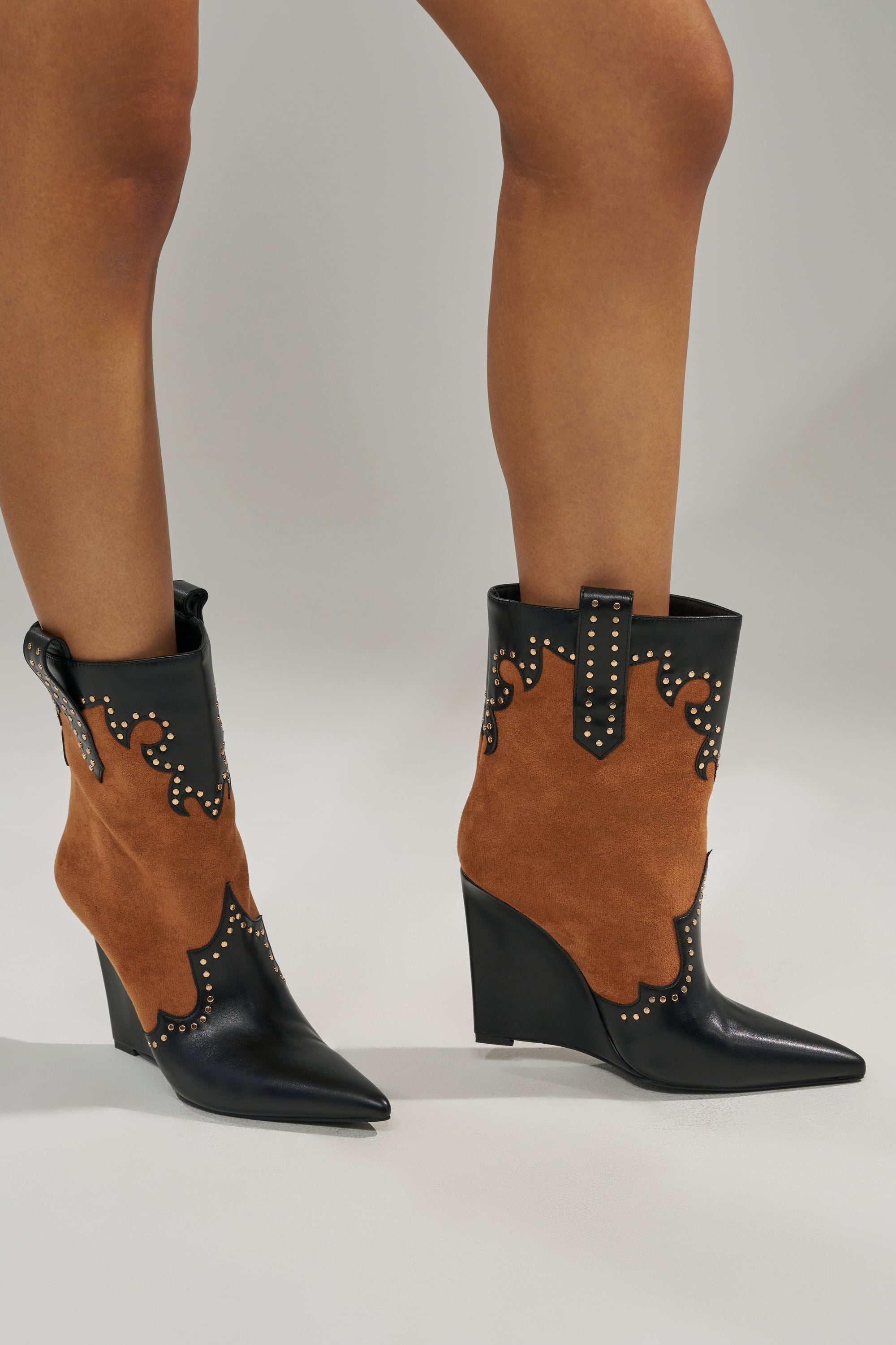 A person wears the AZALEA WANG TALLULAH COGNAC WEDGE WESTERN BOOTIE, featuring black leather, brown cognac suede panels, decorative studs, a pointed toe, and angled wedge heels.