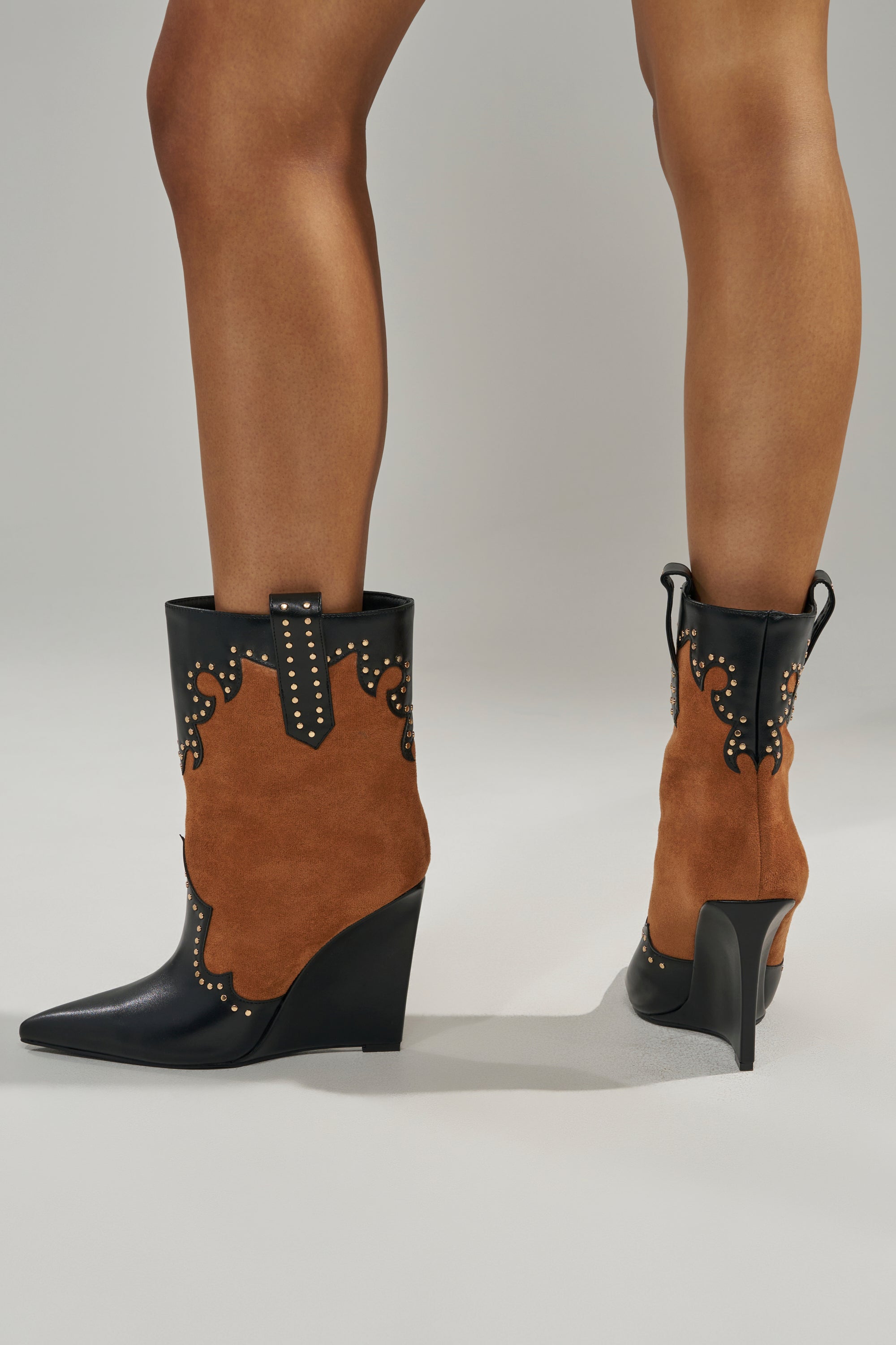 A person stands on a light gray surface wearing the AZALEA WANG TALLULAH COGNAC WEDGE WESTERN BOOTIE, featuring black and cognac suede, pointed toes, decorative studs, and intricate stitching.