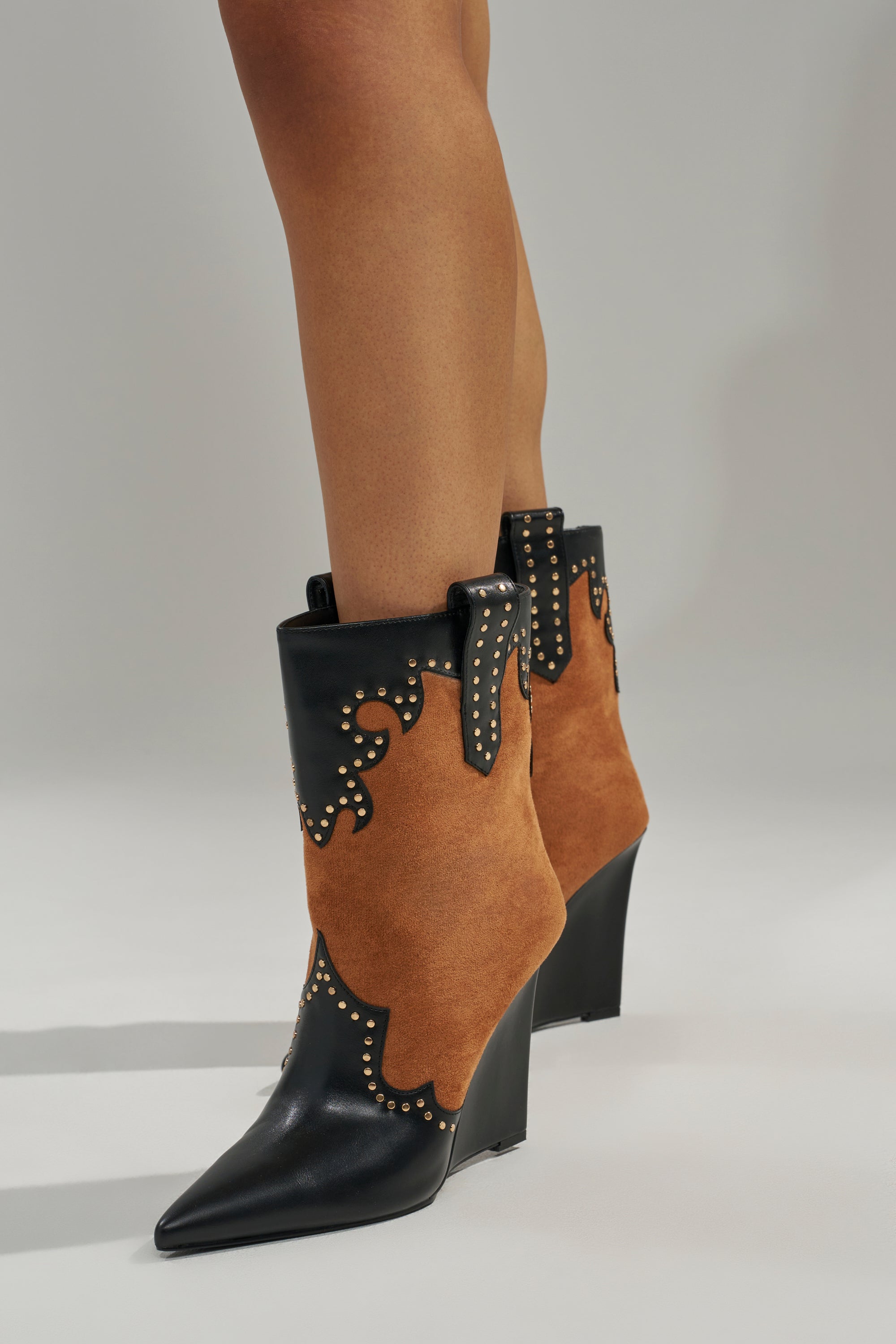 Someone wearing the AZALEA WANG TALLULAH COGNAC WEDGE WESTERN BOOTIE, featuring black leather, brown suede panels, and small gold studs, stands on a light gray surface.