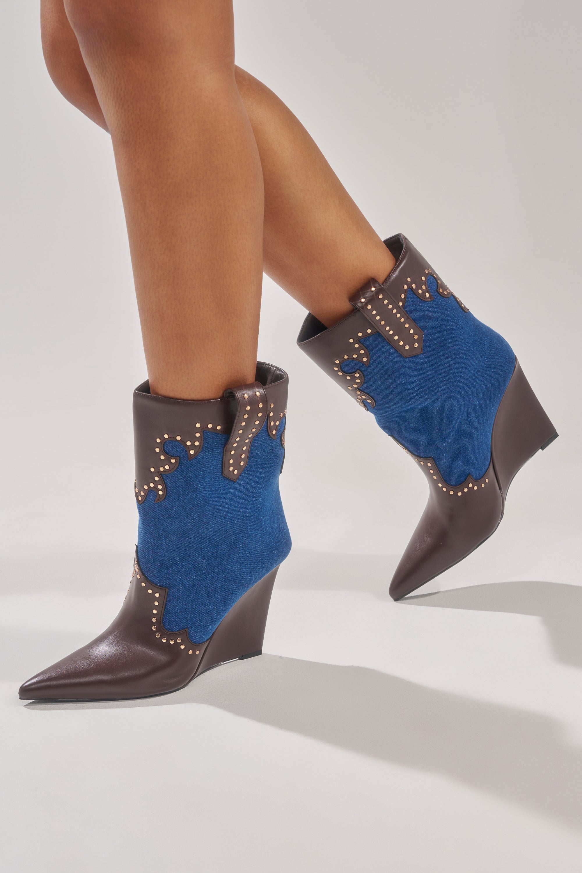Someone stands on tiptoe against a plain background, wearing the AZALEA WANG TALLULAH DENIM WEDGE WESTERN BOOTIE featuring pointed toes, brown leather, blue suede panels, faux leather trim, and gold stud details.