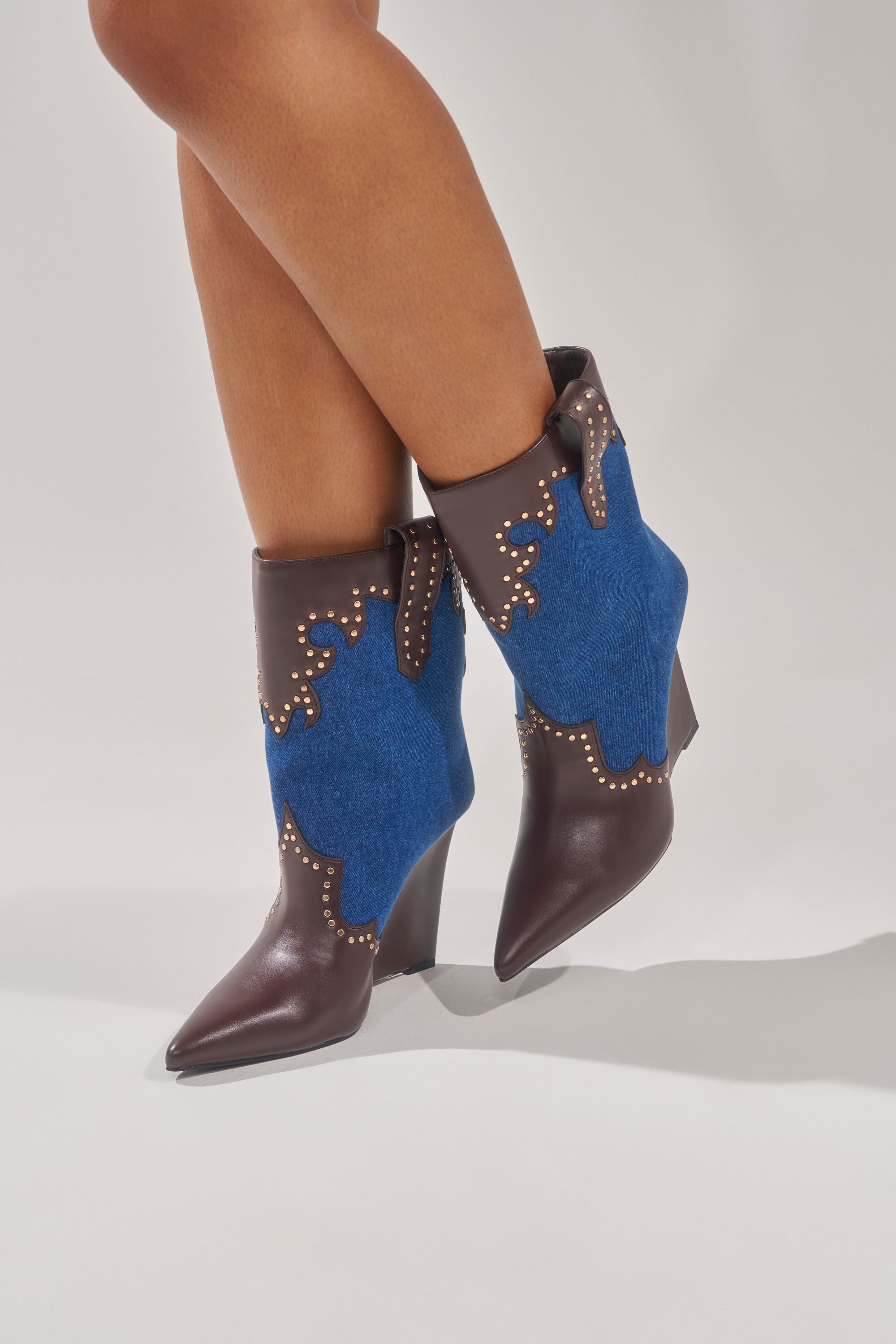 The AZALEA WANG TALLULAH DENIM WEDGE WESTERN BOOTIE features a pointed toe, wedge heel, brown leather and bright blue suede with gold studs, faux leather trim, and decorative western stitching for a bold statement look.