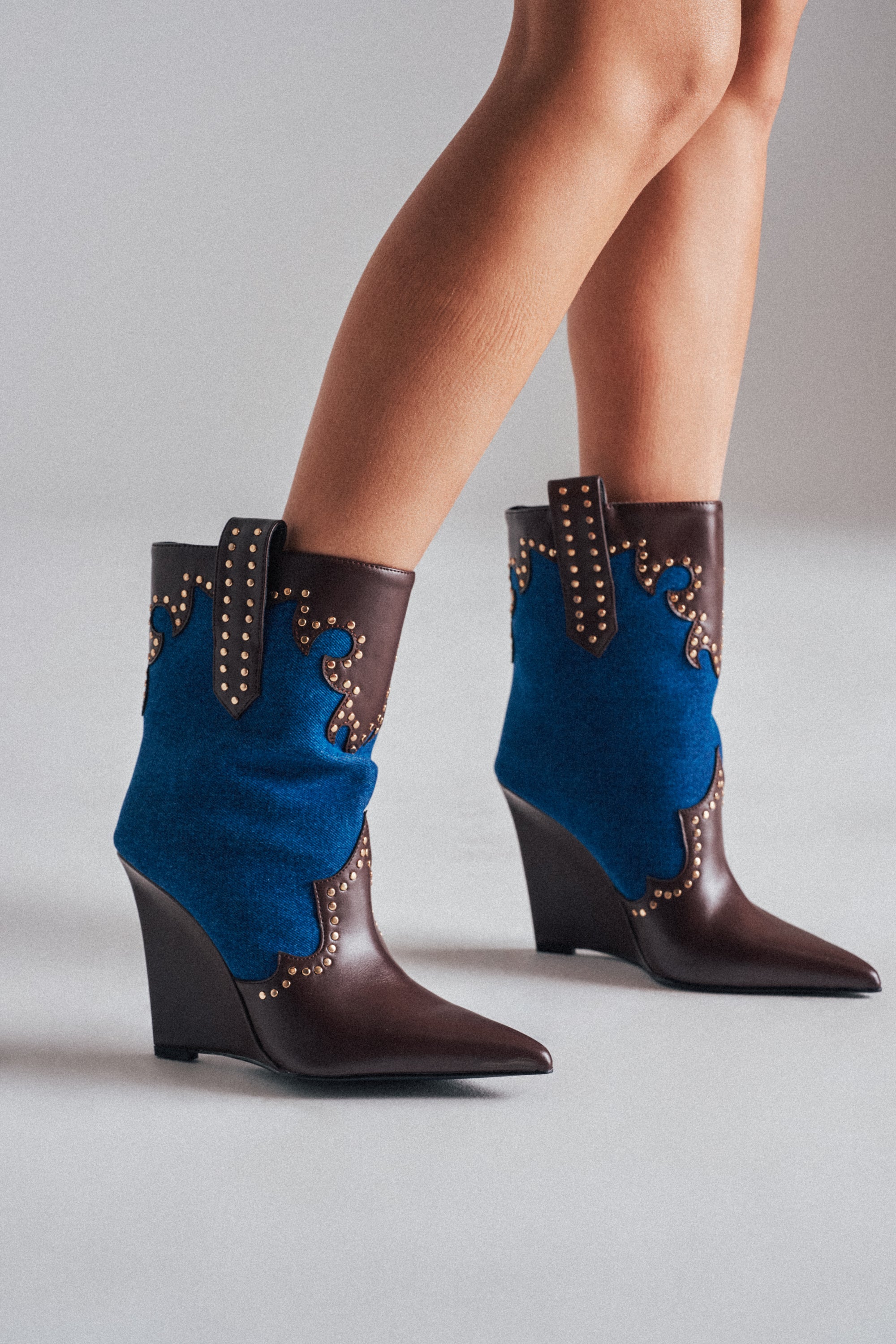 Someone stands on a light gray surface wearing the AZALEA WANG TALLULAH DENIM WEDGE WESTERN BOOTIE, which features brown leather, blue denim accents, pointed toes, and gold decorative studs.