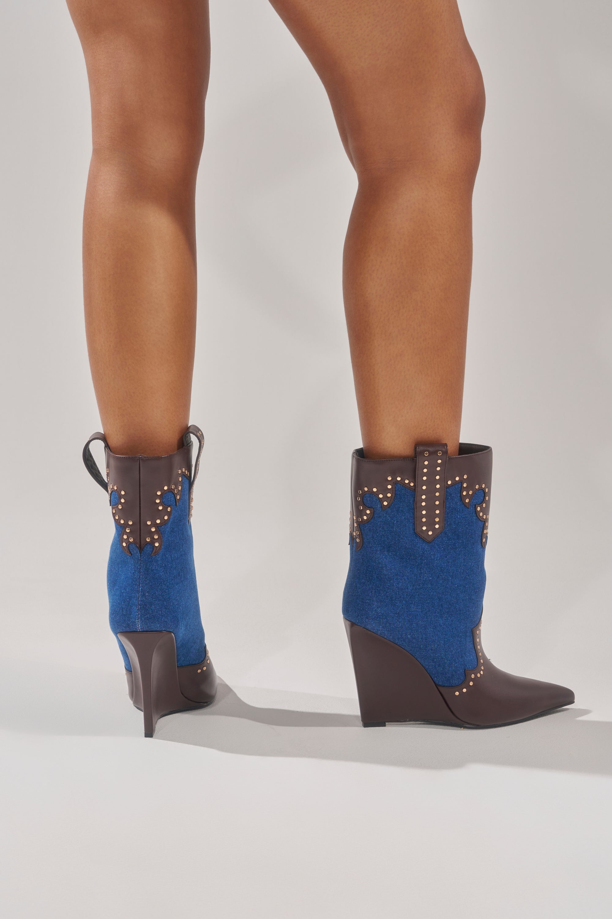 Someone wearing the AZALEA WANG TALLULAH DENIM WEDGE WESTERN BOOTIE, featuring pointed toes and wedge heels, stands against a plain light background.