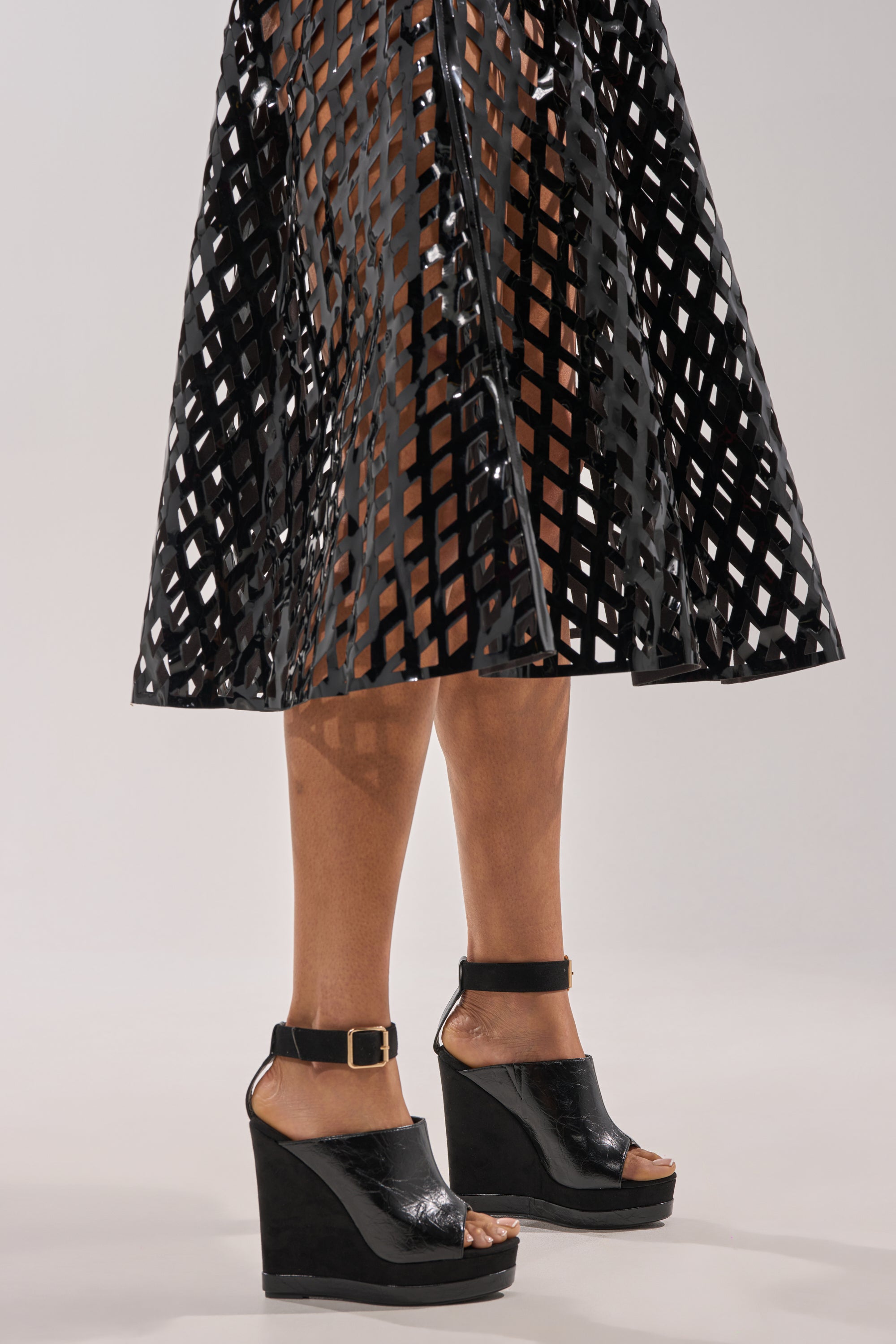 A person stands against a plain light background, wearing a shiny black lattice-patterned skirt and the AZALEA WANG TANA BLACK MIXED MATERIAL WEDGE sandals with ankle straps and a comfort cushion insole. 0