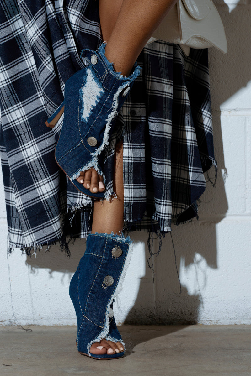 A close-up shows an individual styled with a plaid skirt, featuring a black and white check pattern and frayed hem, against a white brick wall, while wearing AZALEA WANG TANDY DARK DENIM OPEN TOE BOOTIES with stiletto heels, large buttons, and distressed detailing at the edges.