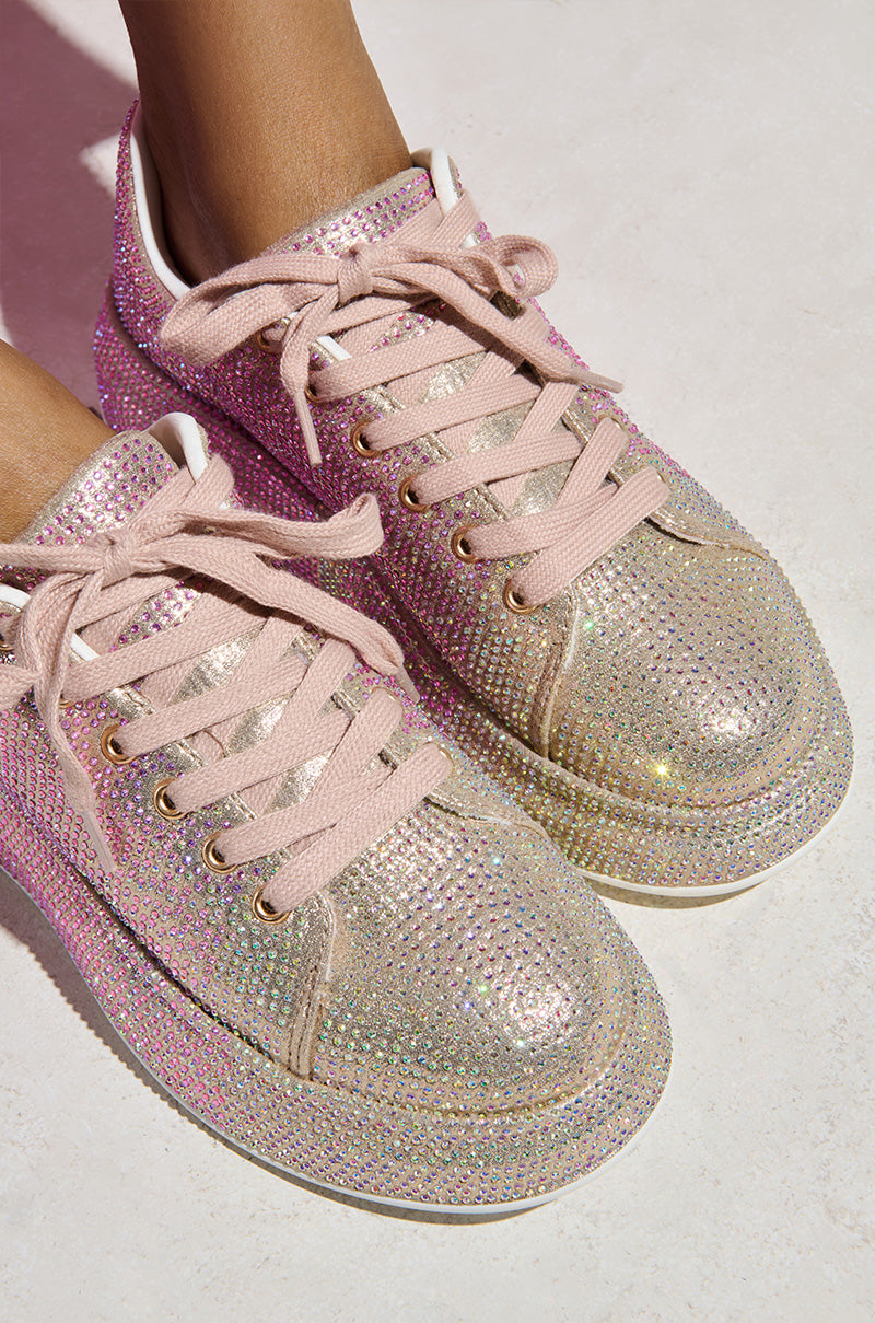 Close-up of someone wearing AZALEA WANG TANGY PINK OMBRE RHINESTONE SNEAKER with glitter textile uppers dazzled by pink and silver rhinestones. Resting on a light-colored surface, the pink-laced shoes create a shimmering effect.
