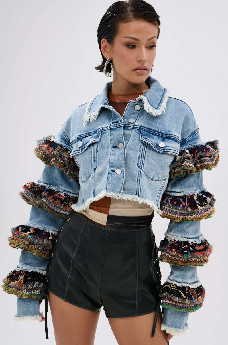 A woman styles the STACKED TAPESTRY DENIM JACKET—with colorful, ruffled sleeves and frayed edges—over high-waisted black leather shorts. Slicked-back hair and large hoop earrings complete her boho-chic look.