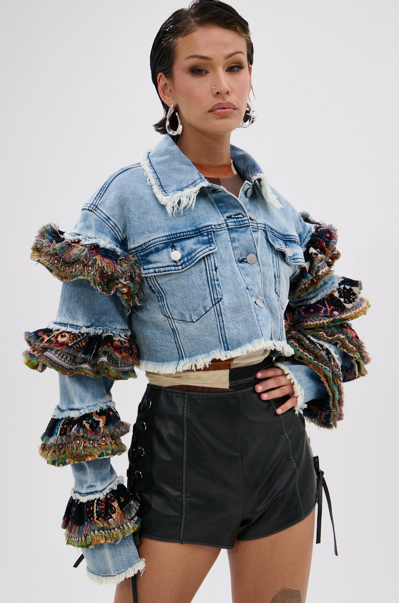 A woman with short dark hair and hoop earrings wears the STACKED TAPESTRY DENIM JACKET, a cropped outerwear piece with frayed edges and ruffled sleeves, paired with high-waisted black leather shorts. Her expression is neutral.