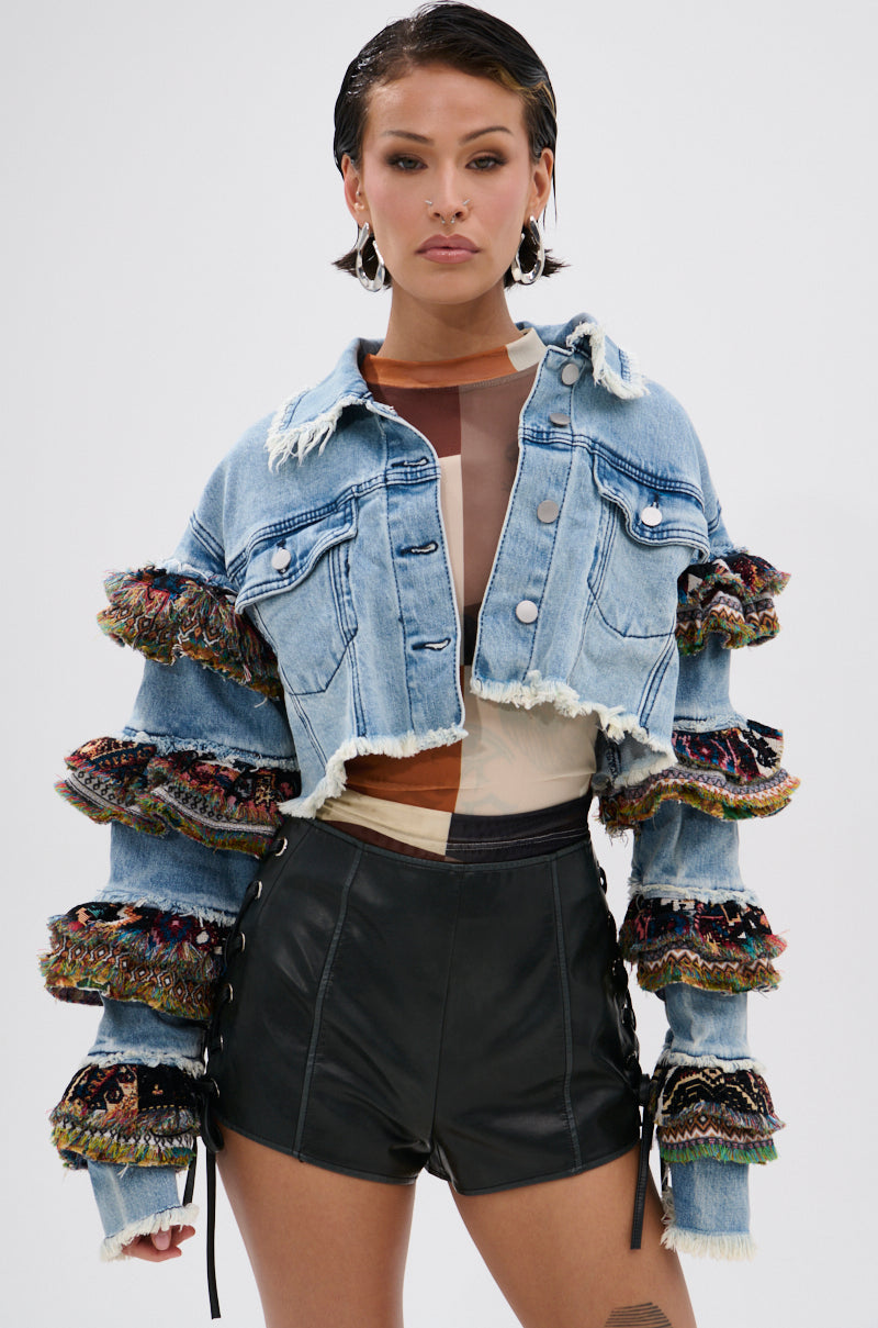 A woman faces forward, wearing the STACKED TAPESTRY DENIM JACKET with frayed edges and colorful embroidered ruffle sleeves, paired with high-waisted black leather shorts and hoop earrings for a boho-chic look.