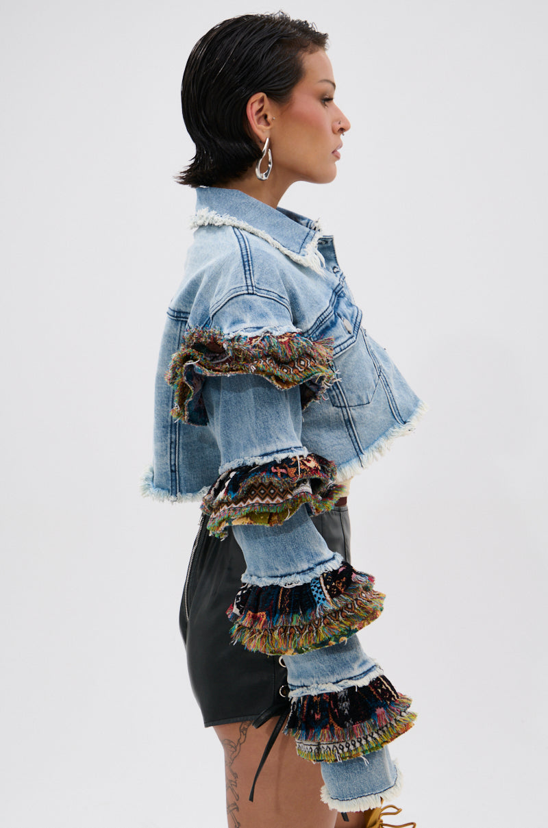 A woman with short dark hair stands in profile, wearing the STACKED TAPESTRY DENIM JACKET with colorful ruffled sleeves, paired with high-waisted black shorts, hoop earrings, and showing a thigh tattoo.