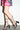 A person wearing a pink and black patterned dress and AZALEA WANG TARANIS Black Curved Chunky Heel Lace Up Sandals stands against a white wall, one foot slightly in front of the other.