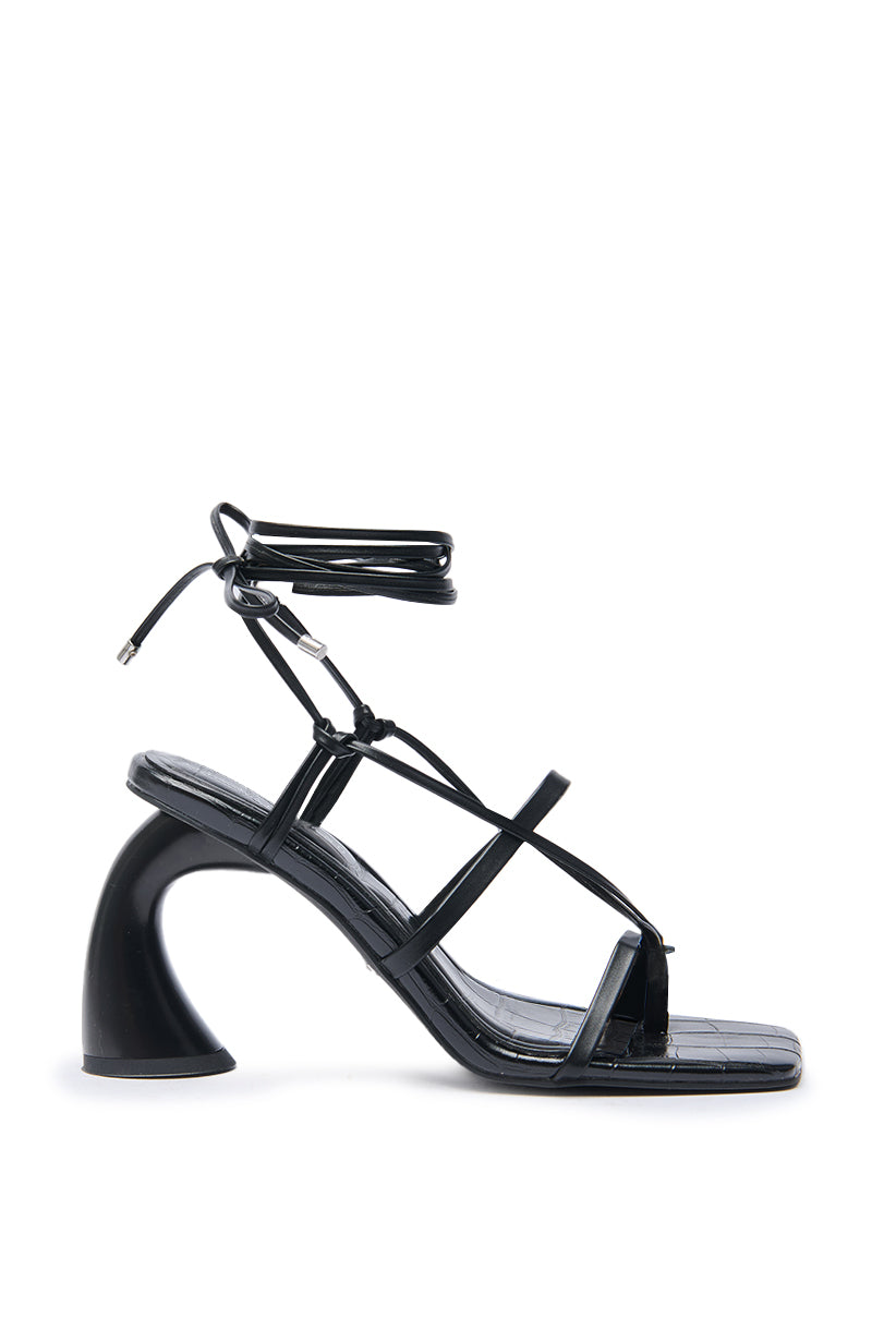 The AZALEA WANG TARANIS Black Curved Chunky Heel Lace Up Sandal features black faux leather, a square toe, lace-up ankle straps, and a sculptural heel, shown in side profile against a white background.