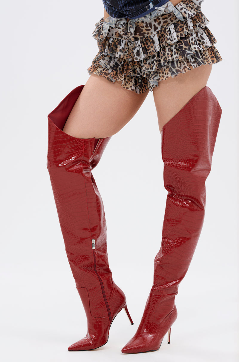 A person wears the AZALEA WANG TARRANT RED STILETO BOOT WITH WESTERN TOPLINE—thigh-high, pointed-toe, crocodile-embossed red boots—paired with ruffled leopard print shorts, standing against a plain white background. 0